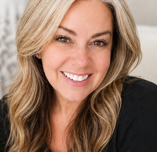 Close-up portrait of a smiling woman with long, wavy blonde hair and bright eyes, wearing a black top.